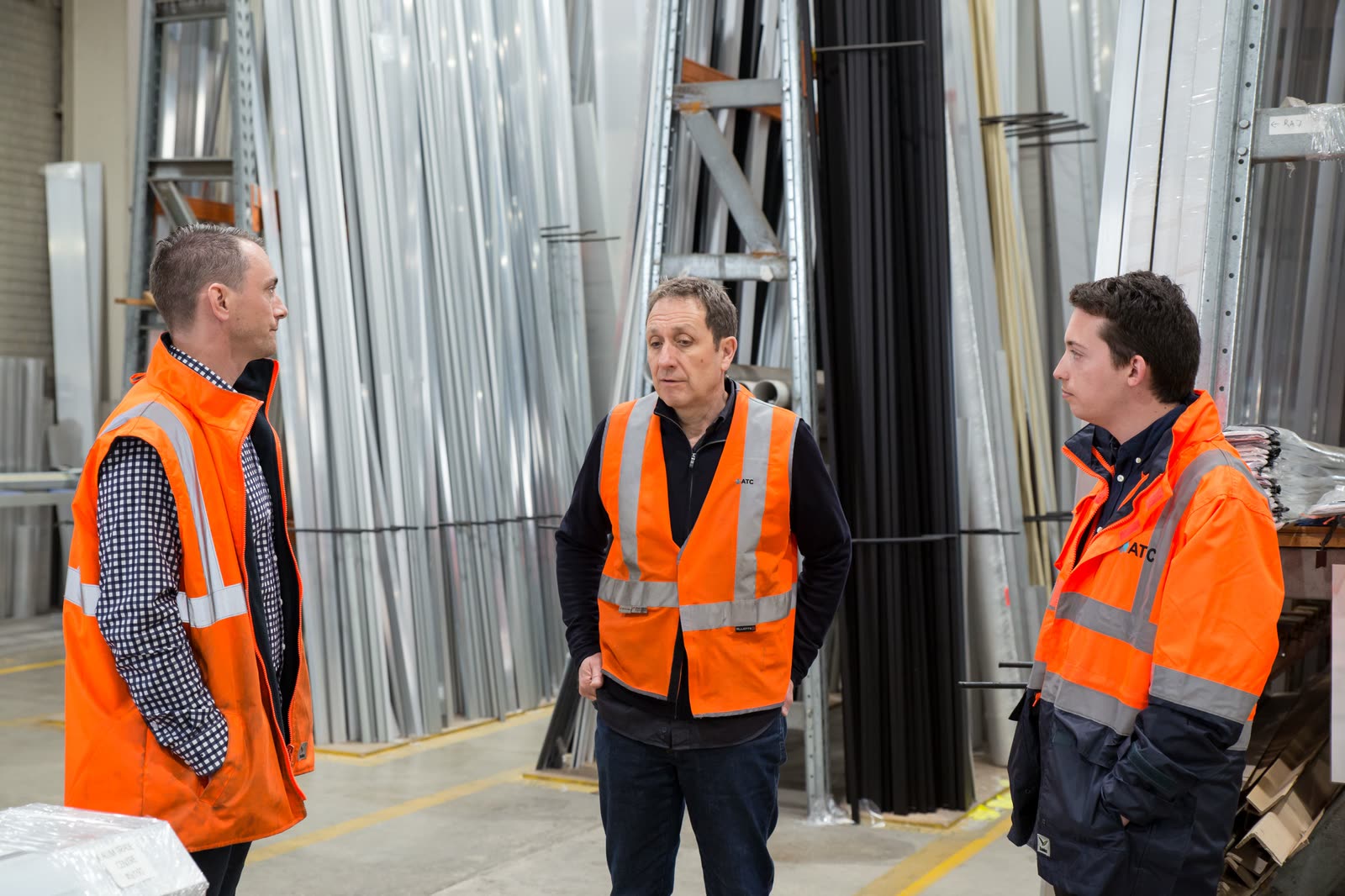 Three people in high-visibility vests talking in a metal-clad workshop.